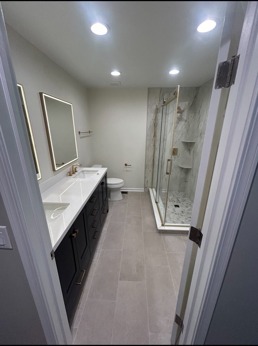 Bathroom remodel with a navy double vanity, backlit LED mirrors and a marble-look glass shower